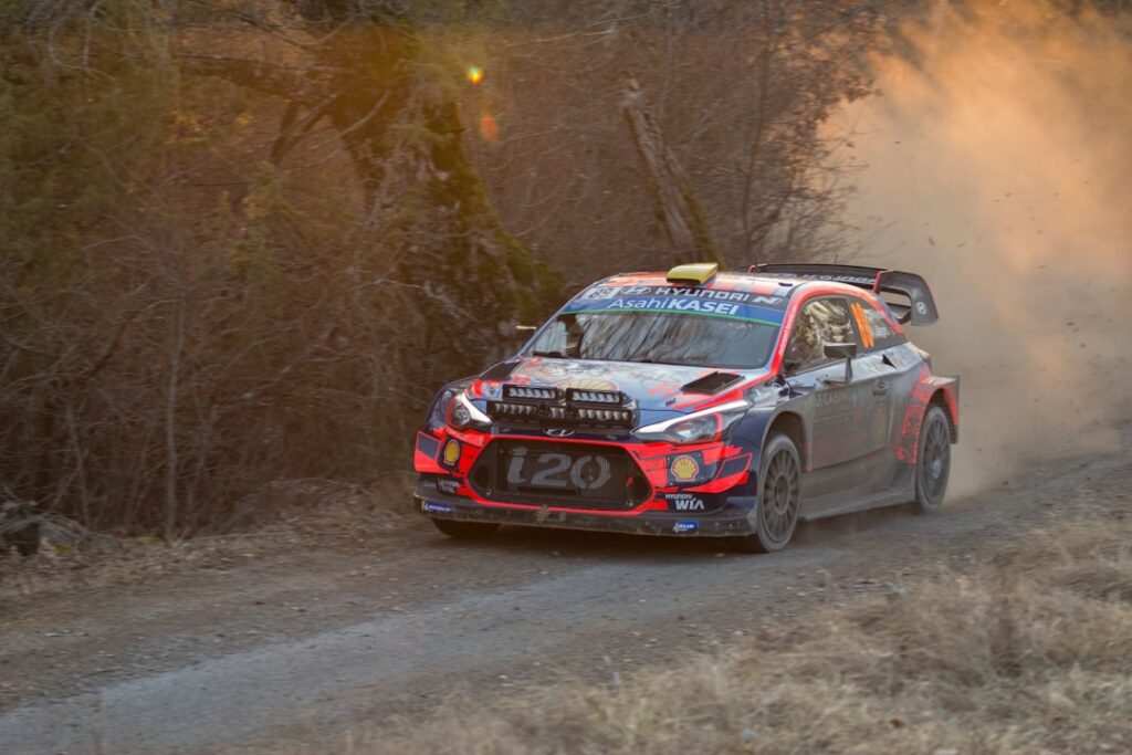 A rally car speeds along a dusty dirt road in rural Argentina with spectators standing close to the course without barriers.